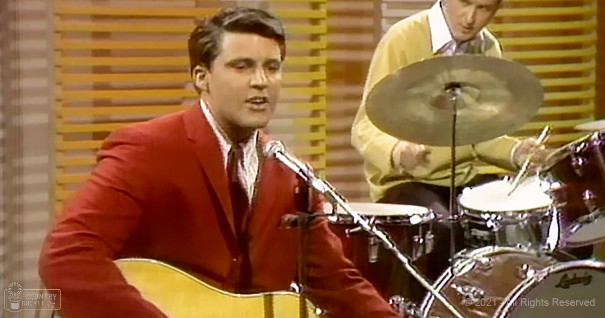 A young adult male singer in a red blazer performs into a microphone while playing guitar, with another adult male drummer in the background wearing a patterned shirt and using a drum set. The setting is an indoor stage performance from the mid-20th century.