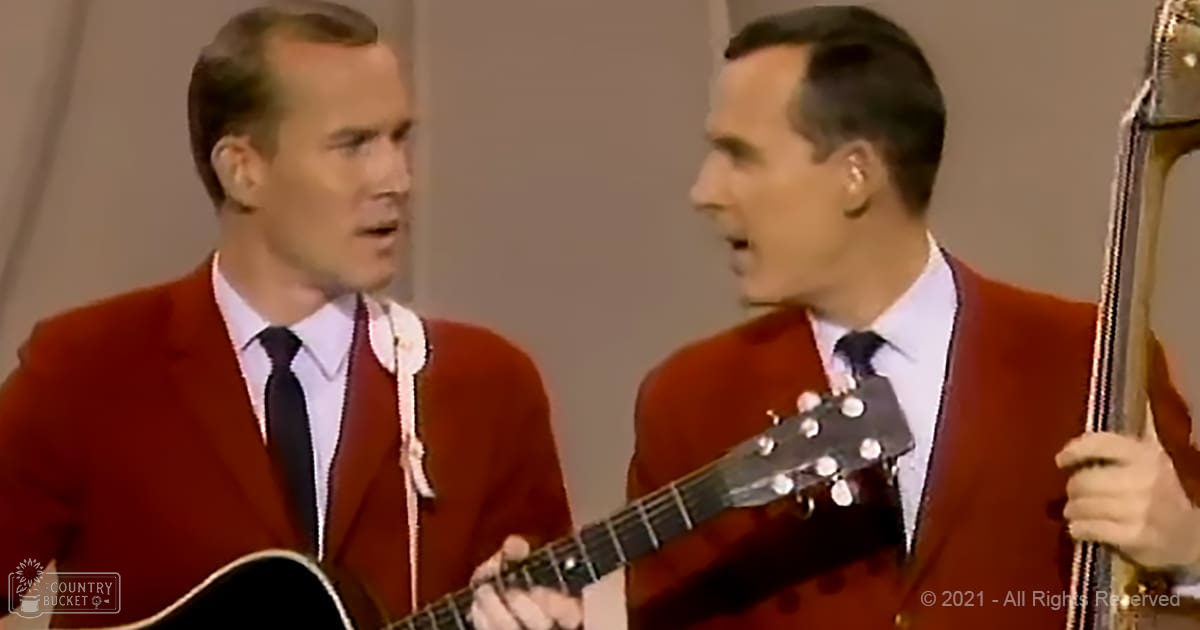 Two adult male singers in a studio setting, wearing red blazers with white shirts and dark ties. A black acoustic guitar is held by the singer on the left; a banjo is partially visible being held by the singer on the right.