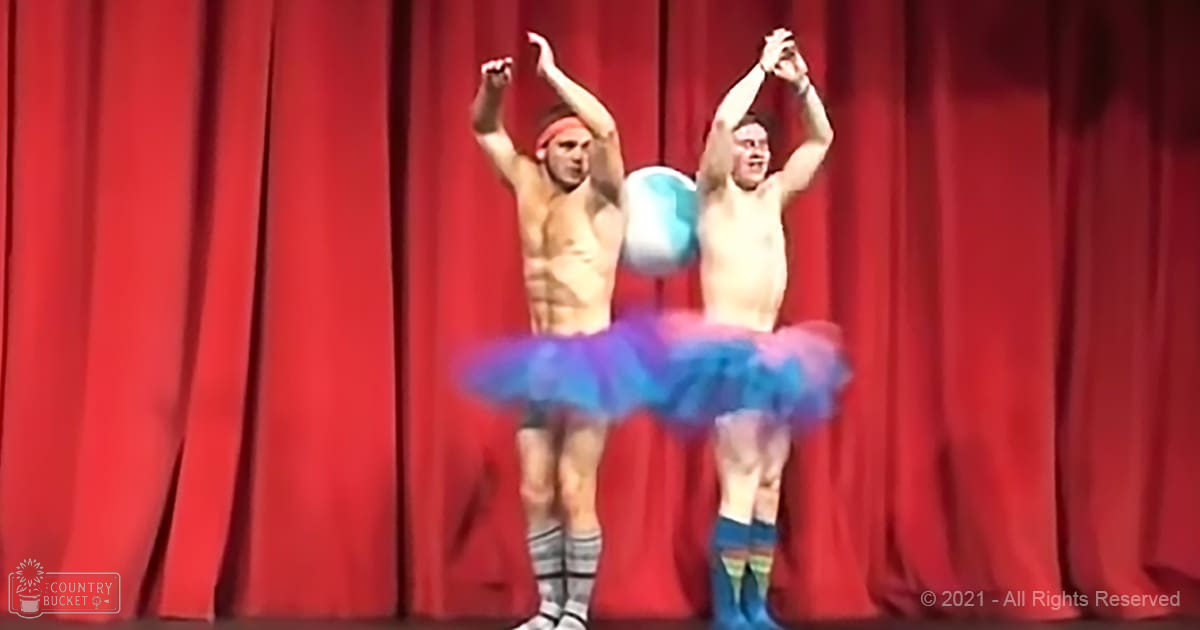 Young adult male performers wearing tulle tutus in shades of blue and purple with ruffled designs, standing on a stage with red curtains as backdrop.