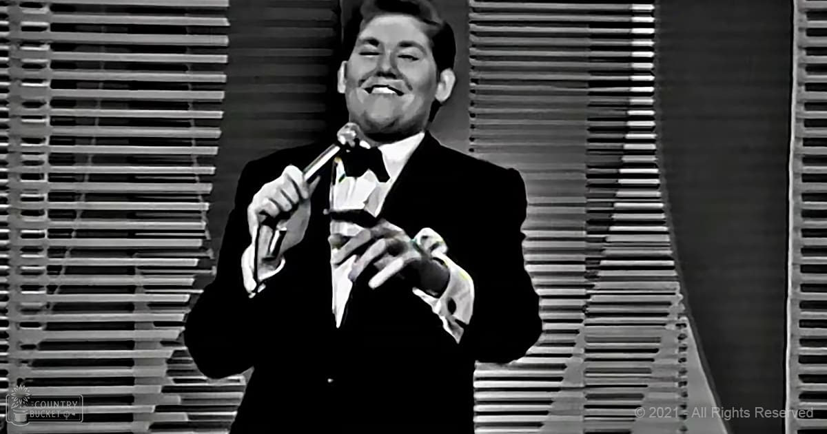 Adult male performer in a black tuxedo with white shirt and bow tie, holding a microphone on stage under bright lighting.