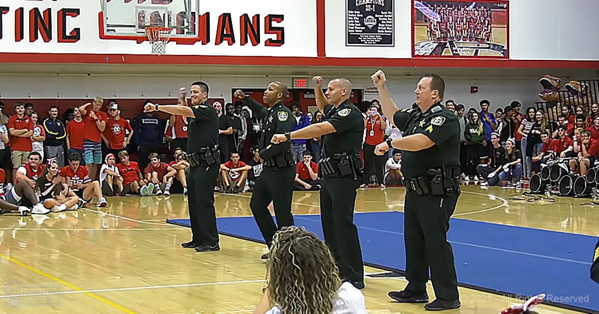 A group of police officers in uniform performing a dance routine in an indoor gymnasium with spectators watching during what appears to be a school or community event.