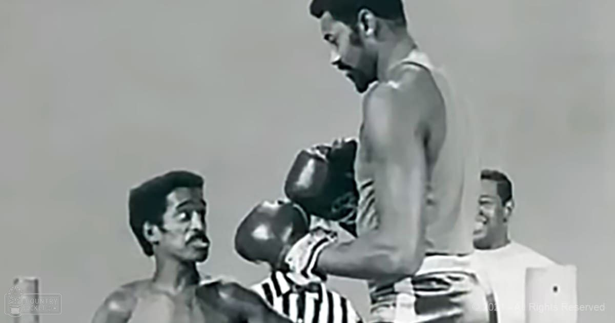 An adult male boxer wearing a white tank top, black shorts, and black gloves, surrounded by other adults in an indoor boxing ring setting with neutral-colored walls and bright lighting.
