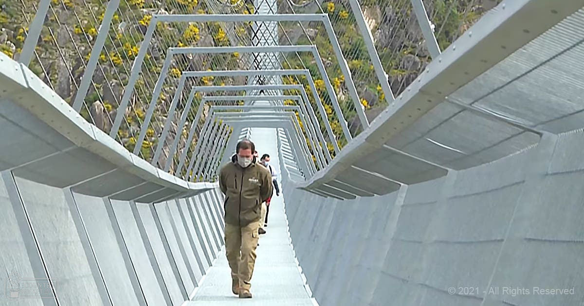 An adult male pedestrian wearing a green jacket and beige pants crosses an outdoor metal bridge with another person in white shirt and dark pants, surrounded by greenery and yellow flowers under natural daylight.