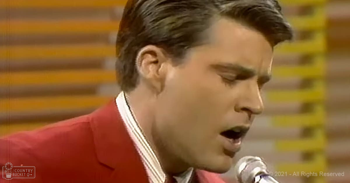A male adult in a mid-20th-century red jacket with a white shirt, singing into a microphone in an indoor studio setting.
