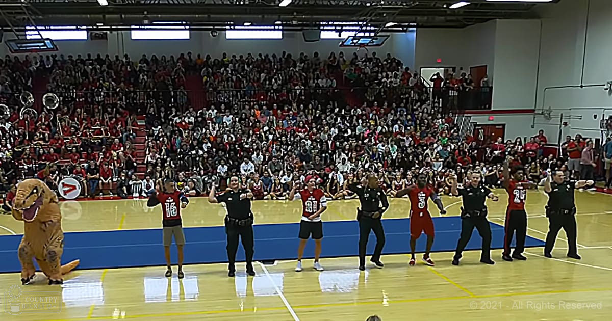 A child in a red and black jersey stands on an indoor basketball court alongside other performers, including a man dressed as a dinosaur. The scene is set in a crowded arena with spectators and artificial lighting.