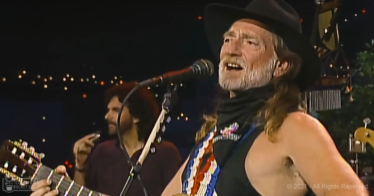 An older adult male singer performs shirtless into a microphone at an outdoor concert venue, wearing a black tank top and dark hat with visible tattoos. A beaded necklace is also prominent.