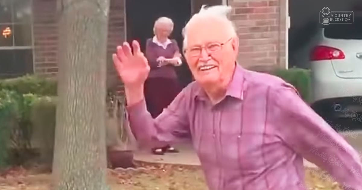 An older adult male waving in front of a brick house with an older adult female inside, wearing glasses and a long-sleeved purple shirt; the scene is outdoors, likely autumn, with soft lighting and greenery around.