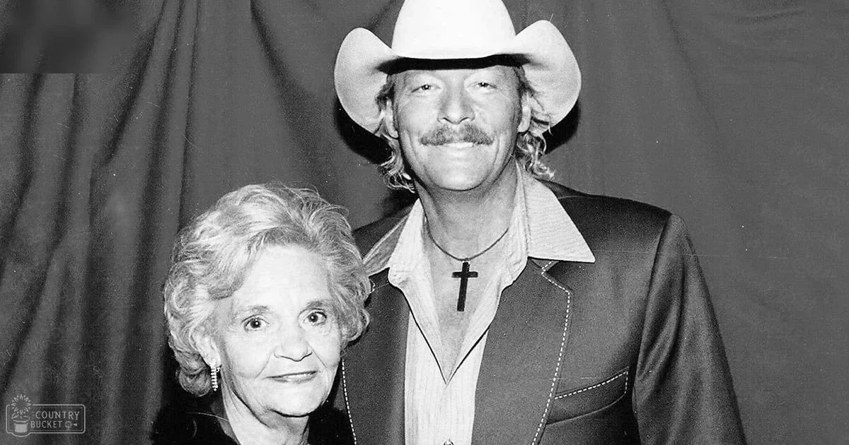 A man wearing a cowboy hat and collared shirt with a cross necklace stands next to an older woman in a dark jacket or coat, both seemingly from the 1970s. The background is a plain curtain indicating an indoor setting.