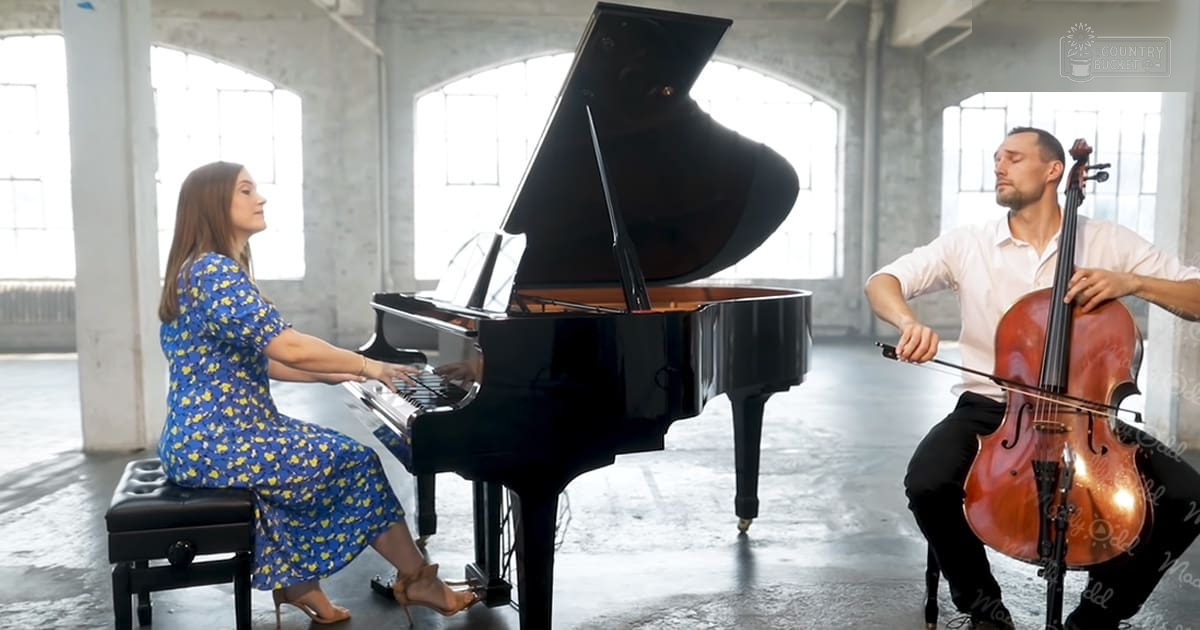 Industrial-style performance space with arched windows, concrete pillars, exposed brick walls; young adult female in blue floral dress plays piano, adult male in white shirt and black pants plays cello under natural light.