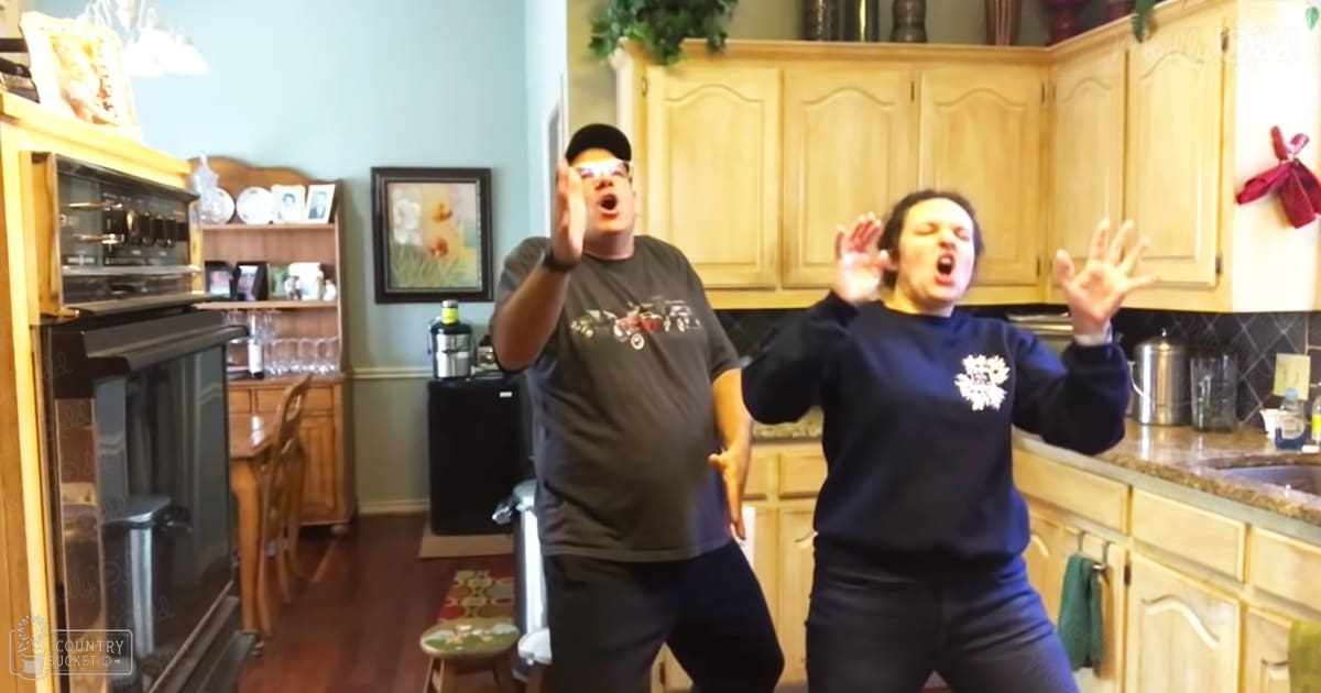 Two adults, a male in a dark gray t-shirt with a graphic print and black pants, wearing a black watch, standing next to an adult female in a dark blue sweatshirt with white text and dark pants, in a well-used residential kitchen with wooden cabinets, a small black refrigerator, and various kitchen utensils. The setting is indoors with bright lighting.