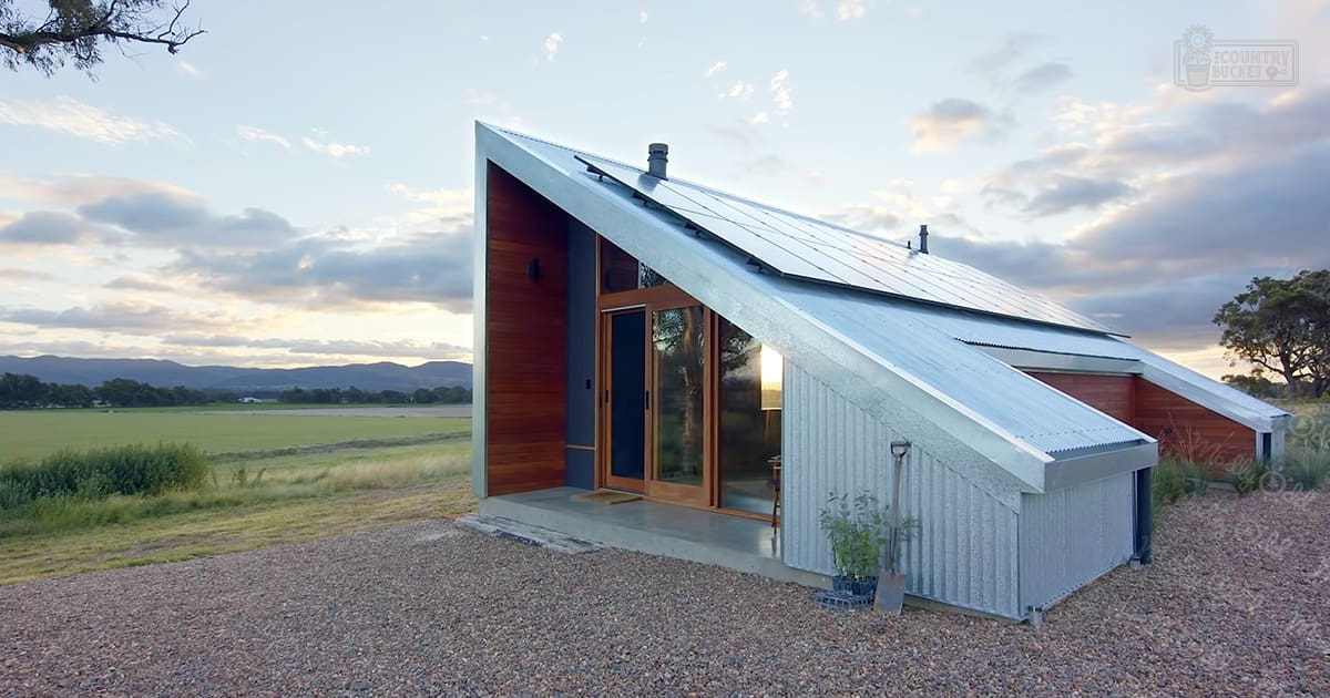 A rural modern eco-friendly house with solar panels, gardening tools, and large glass windows set against open fields under a soft evening sky.