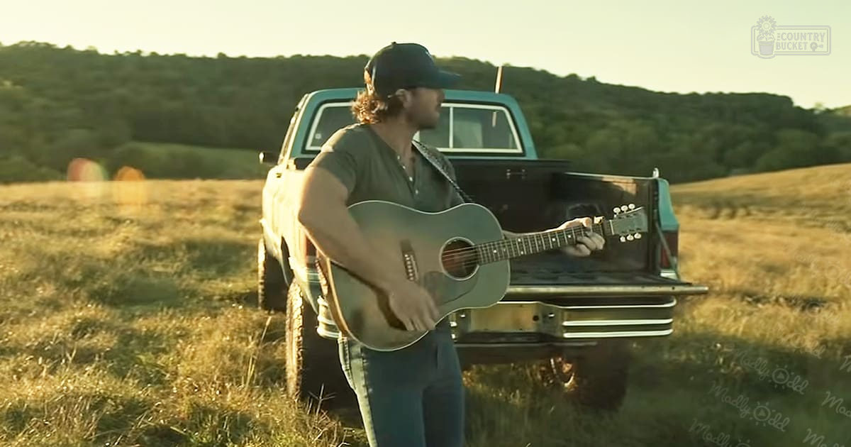 Adult male playing guitar in an open field with a vintage pickup truck, late spring or summer, rolling green hills, warm golden light casting long shadows.