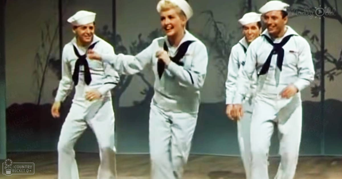 A group of performers in a theatrical setting, dressed in white sailor suits with dark blue neckerchiefs and caps, standing before a stage backdrop with foliage and architectural elements.