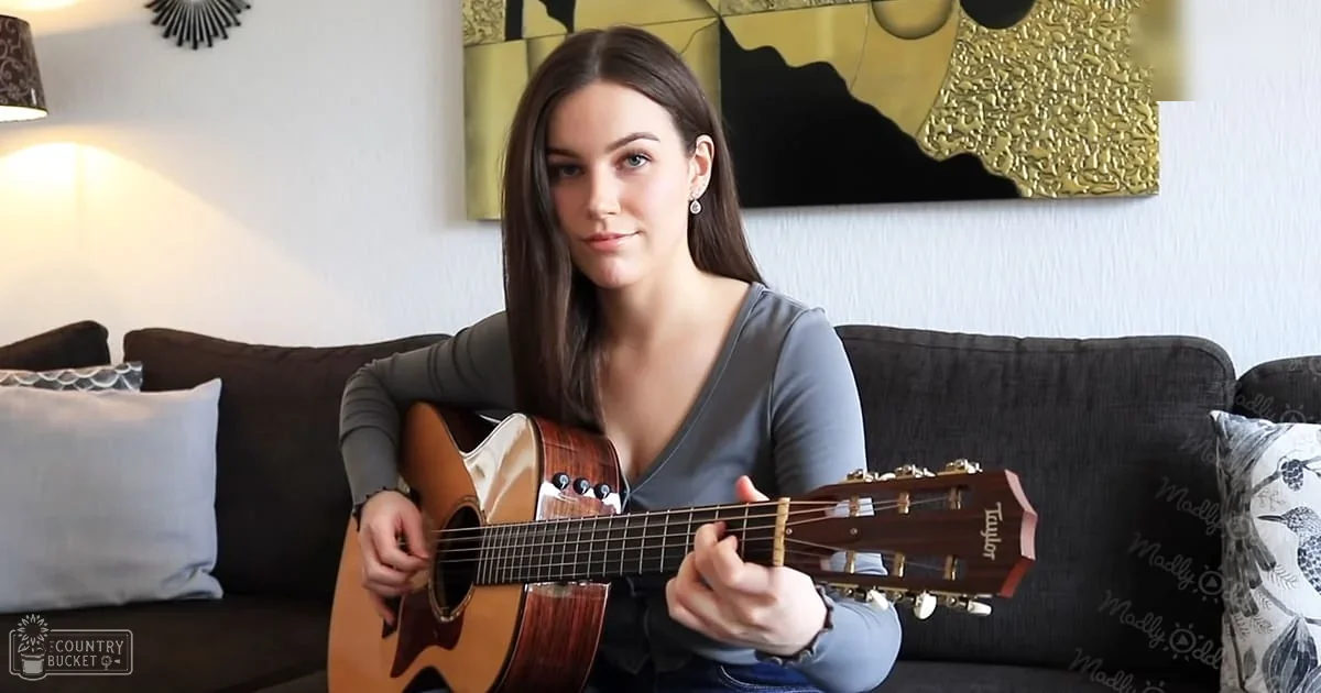Young adult female playing guitar in a living room with gray top and blue jeans, surrounded by furniture and home decor.