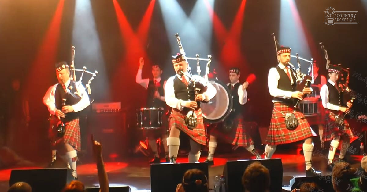 A Scottish pipe band performing under red and white spotlights, dressed in traditional attire with a red plaid kilt and dark vest, on an indoor stage setup.