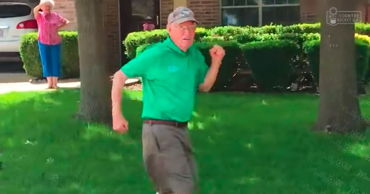 An older adult male wearing a green polo shirt, gray pants, and a backward gray baseball cap stands amidst well-maintained suburban grass with trees and shrubs, while an older adult female in a pink top and blue pants holds her hair back, barefoot, under clear daylight.