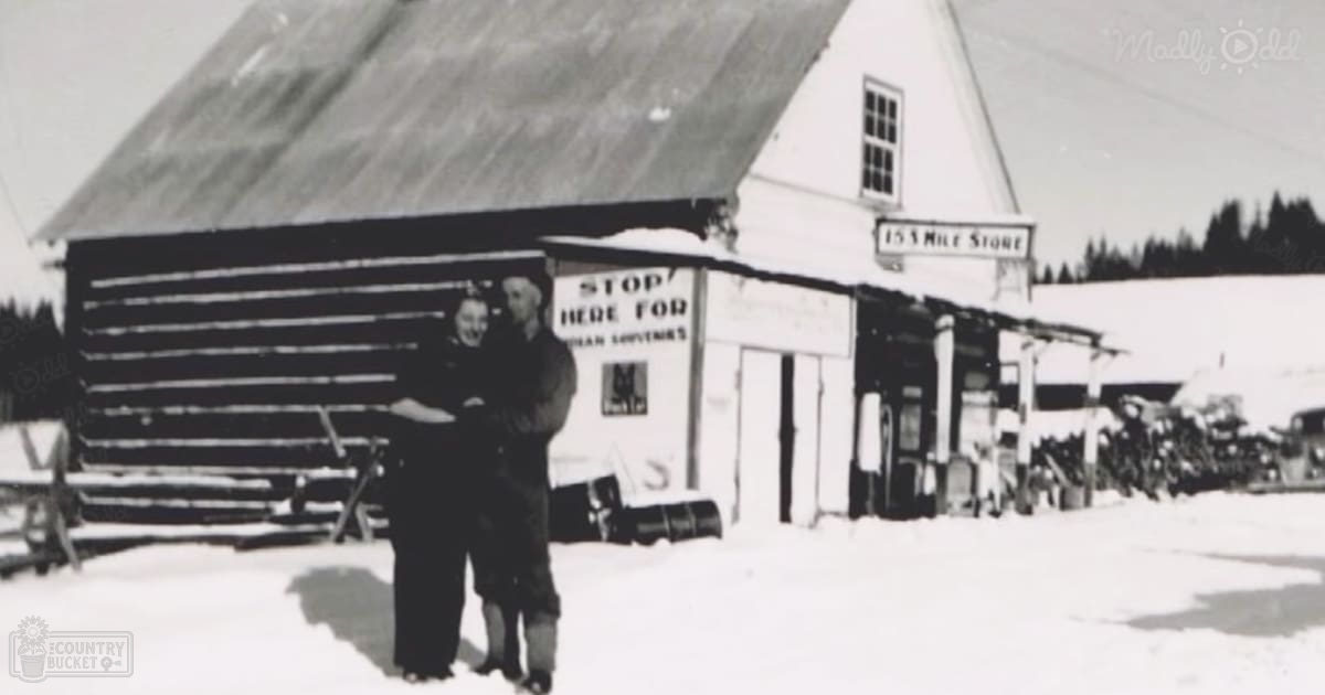 An adult male singer and a child wearing winter attire stand before 'The 15 Mile Store' in Alaska; snow-covered ground and clear daylight highlight the scene.