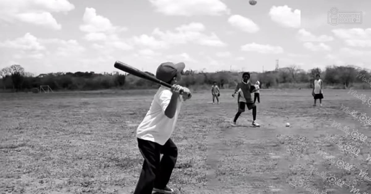 A young adult male child plays baseball in an open field with two middle-aged adults nearby under partly cloudy skies.