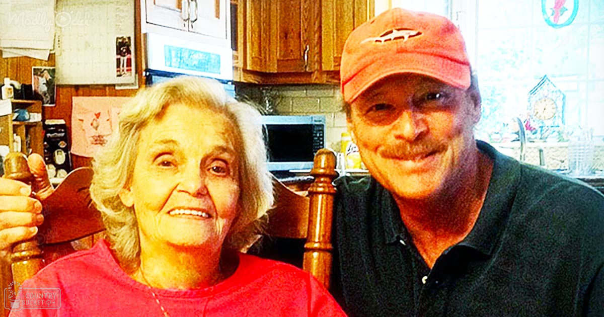 An older adult woman wearing a red t-shirt stands in a well-lit kitchen setting, accompanied by a middle-aged man in a black polo shirt and orange baseball cap; wooden chairs and cabinets are visible with decorative home decor.