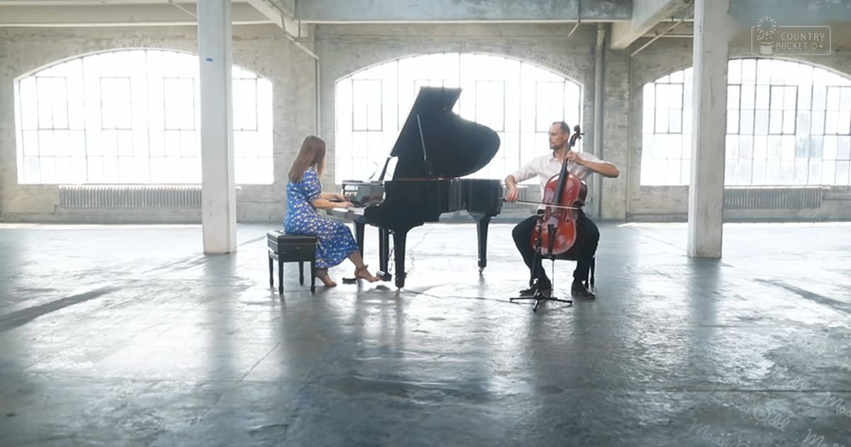 A young adult female pianist in a blue dress performs alongside a middle-aged male cellist in an industrial-style space with large arched windows allowing natural light, suggesting daytime during spring or summer.