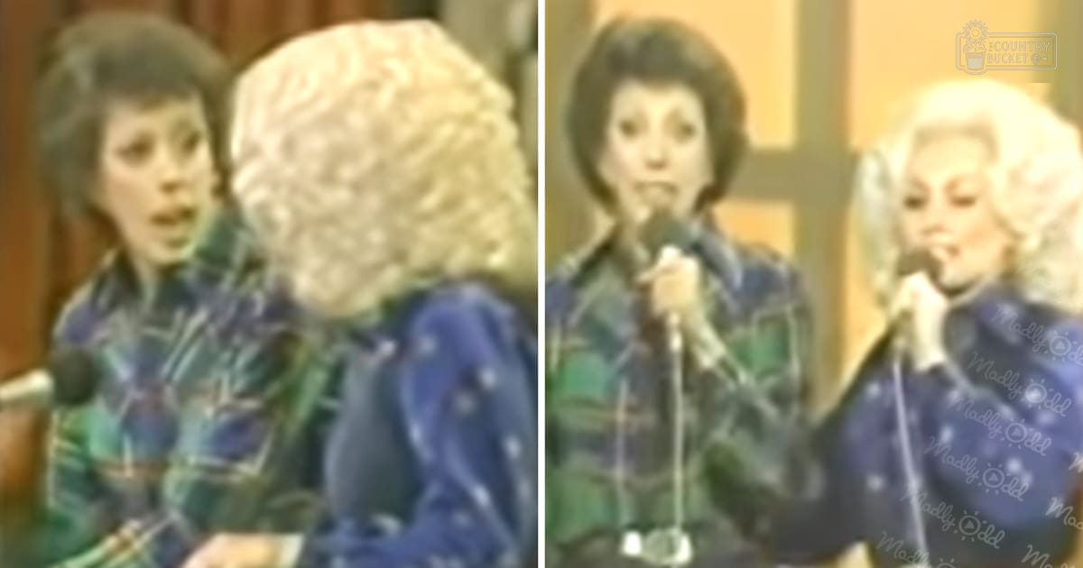 A toddler girl wearing a plaid shirt and dark pants indoors, with an older adult female in a polka dot jacket and scarf standing nearby; indoor setting with wooden paneling and natural light suggesting daytime.