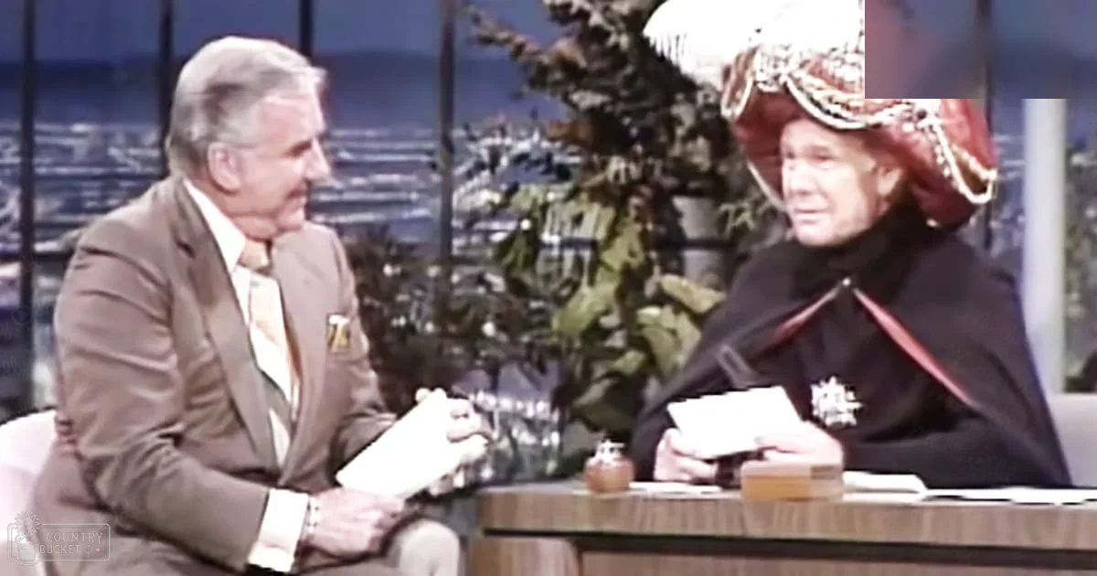 Two older adult males seated in a talk show setting with wooden furniture and large windows showing an outdoor scene of trees and hills.