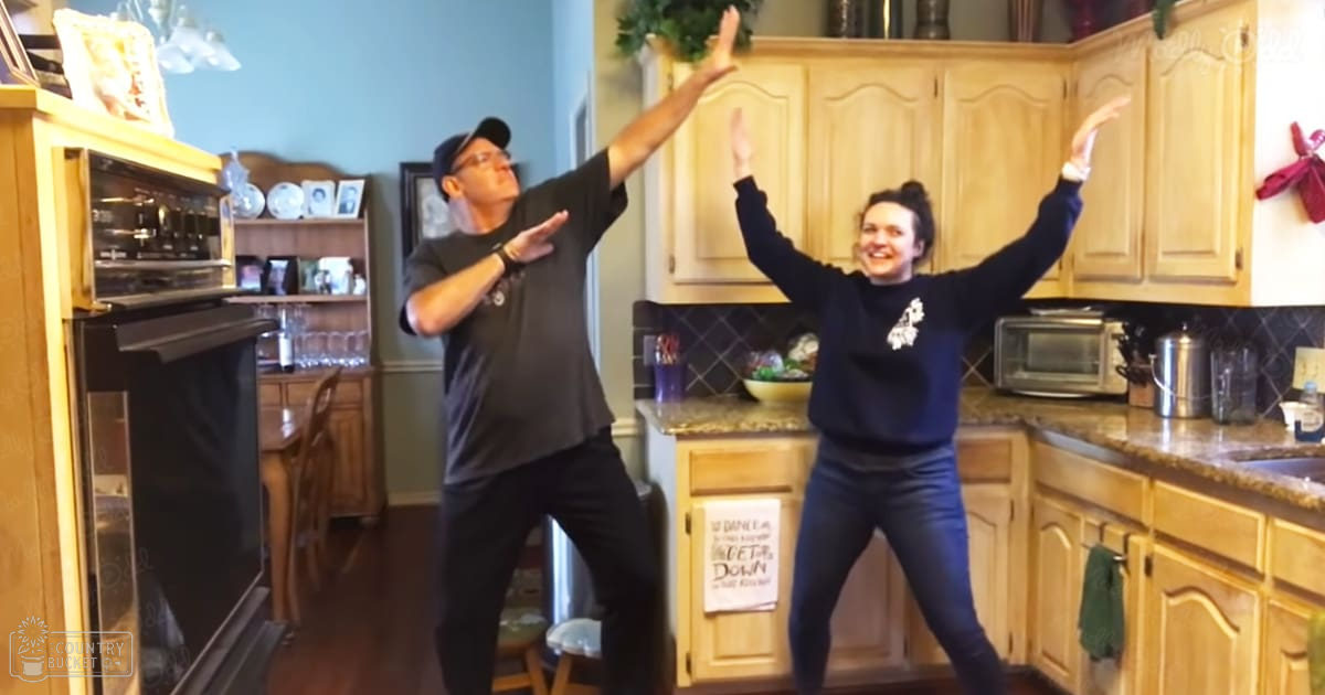 A couple dancing in their kitchen with wooden cabinets and a sign reading 'DANCE LET'S DOWNTOWN'.