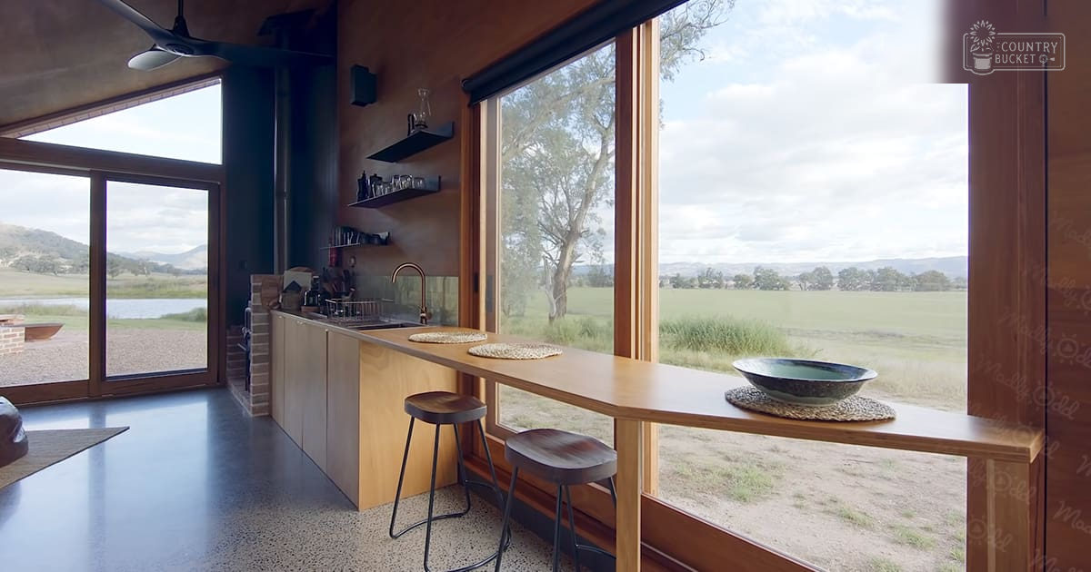 Modern kitchen with large windows overlooking scenic green fields and distant hills under partly cloudy sky, indoor setting with lush vegetation visible through windows, midday natural light, spring or summer season.