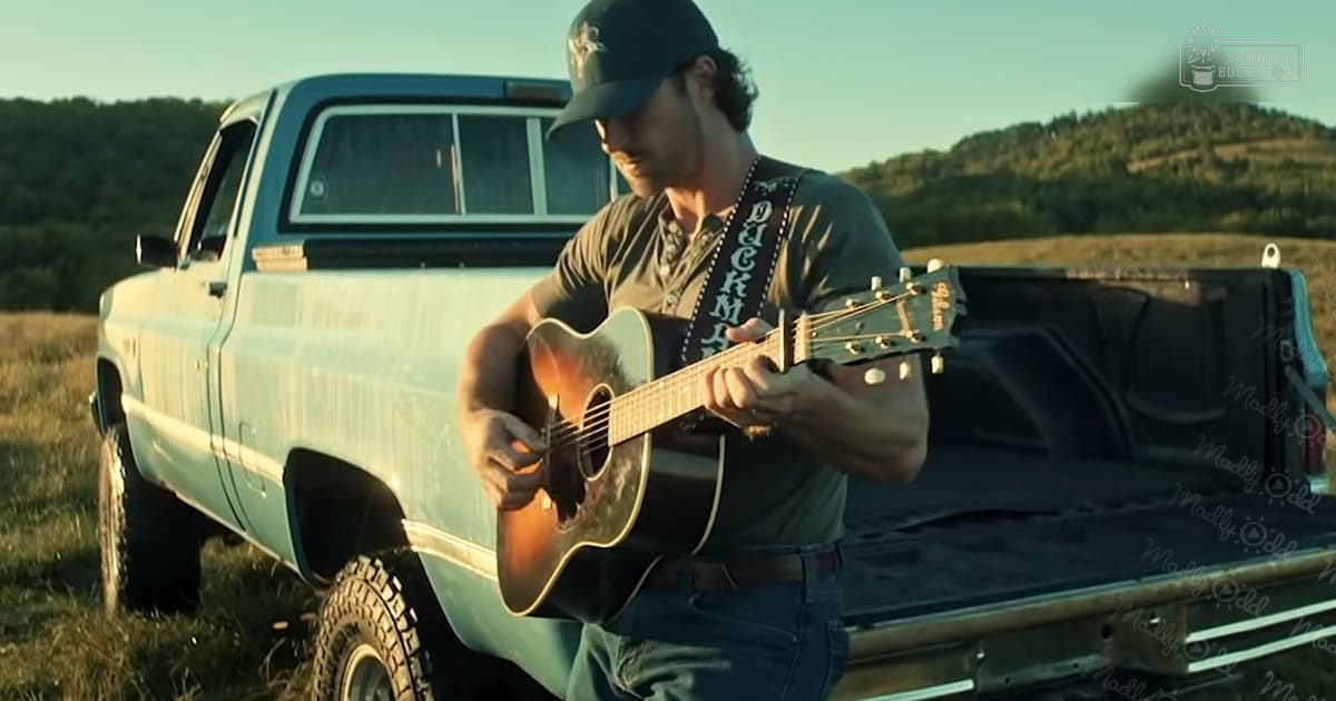 A young adult male in a rural setting plays guitar in front of an old light blue pickup truck, with dry golden grass and hills under warm late afternoon sun.