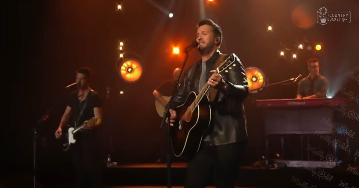 An adult male singer wearing a black leather jacket and dark pants, holding an acoustic guitar in what appears to be a dimly lit studio or stage setting with warm orange lighting.