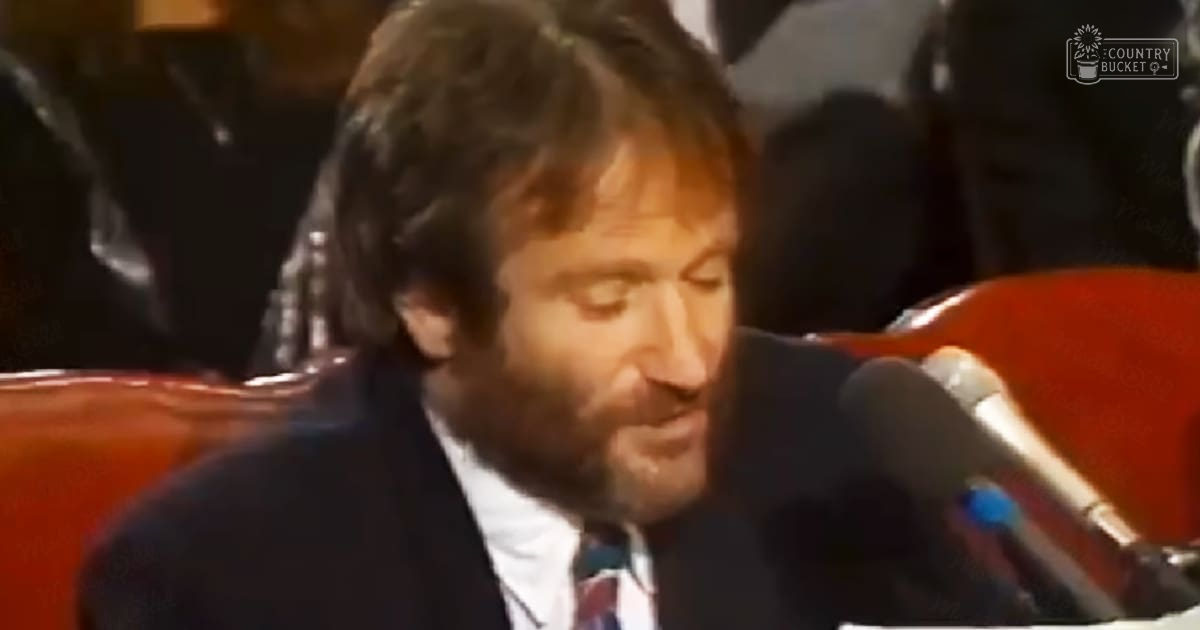 A middle-aged man in a dark suit jacket over a white collared shirt and patterned tie sits indoors, possibly at a formal event or interview setting.