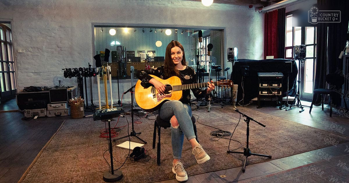 Young adult female in a music studio holding an acoustic guitar, wearing black floral top, blue jeans with rip at knee, beige sneakers. Music recording equipment and natural light suggest daytime scene.