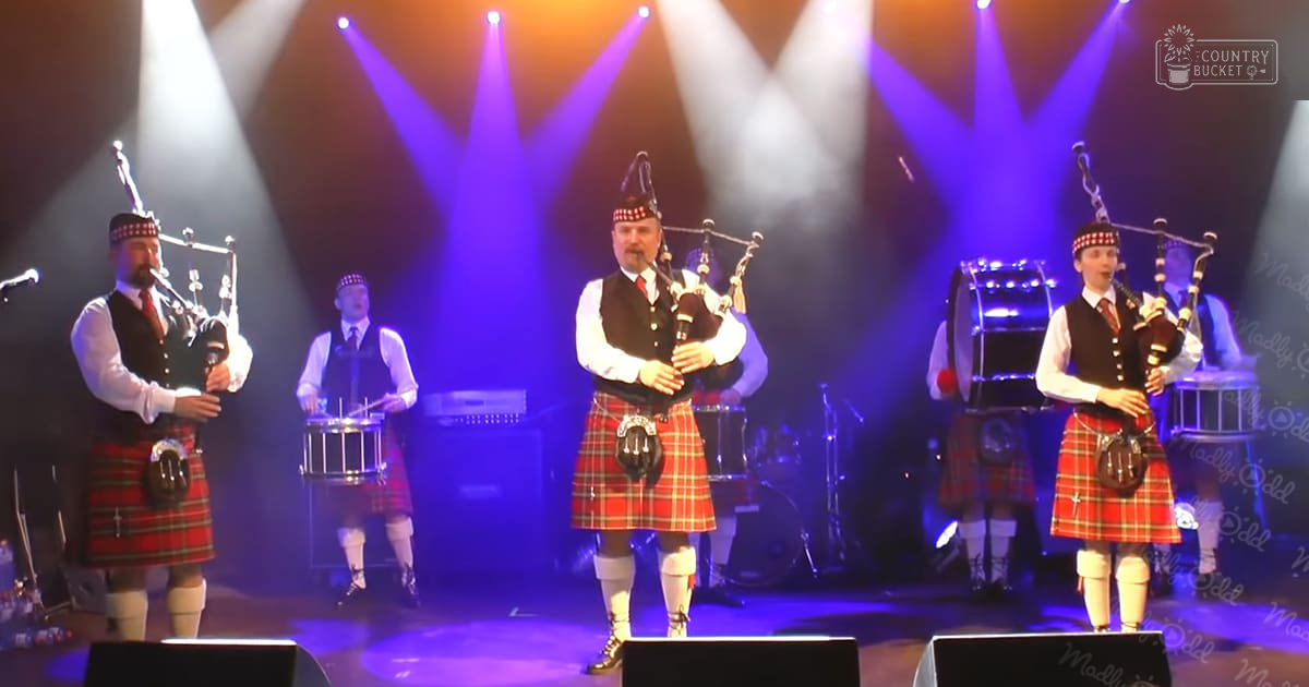 Scottish pipe band performing at indoor concert venue with red tartan kilts under stage lighting.