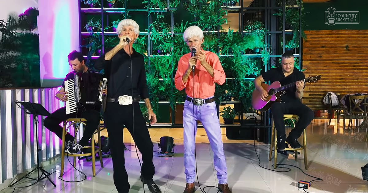 Musical performance in an indoor setting with adult males wearing various clothing. The scene includes chairs for musicians, a stool, sheet music on a stand, plants, and wooden panel design elements.