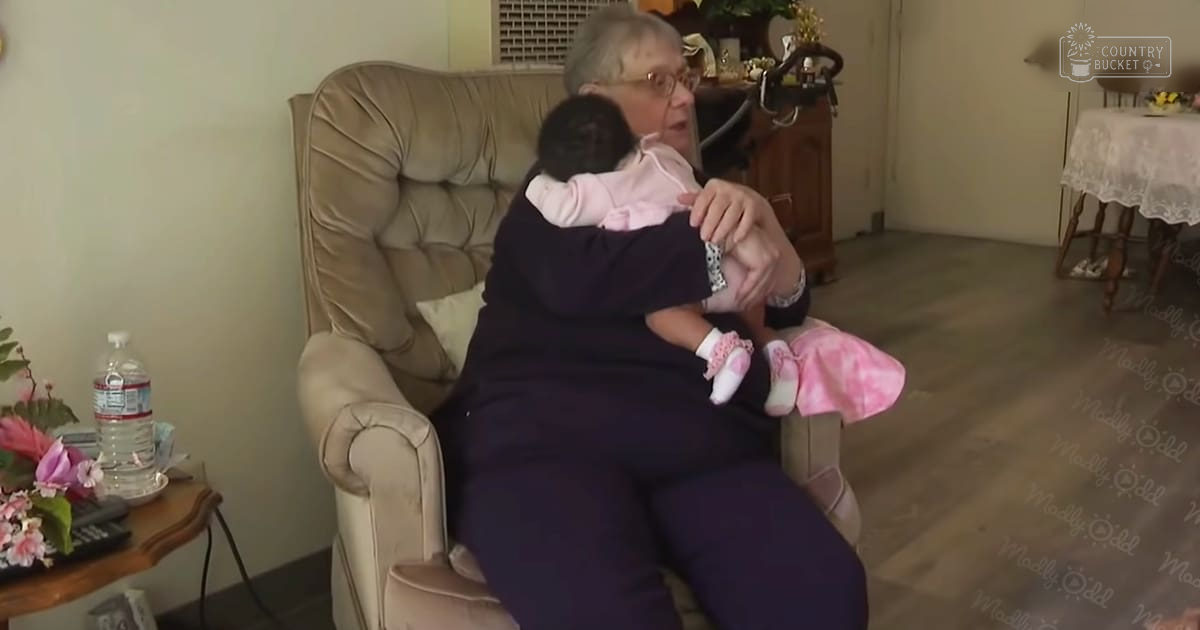 An older adult female wearing dark-colored stretchy pants, glasses, and a watch sits in a beige recliner chair with a baby doll beside her. The cozy living room has wooden furniture, flowers, and natural light.
