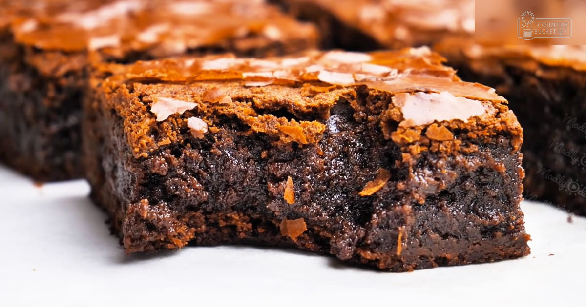A rich, glossy chocolate brownie topped with chocolate shavings and crystalline pieces on a plain white baking surface indoors.
