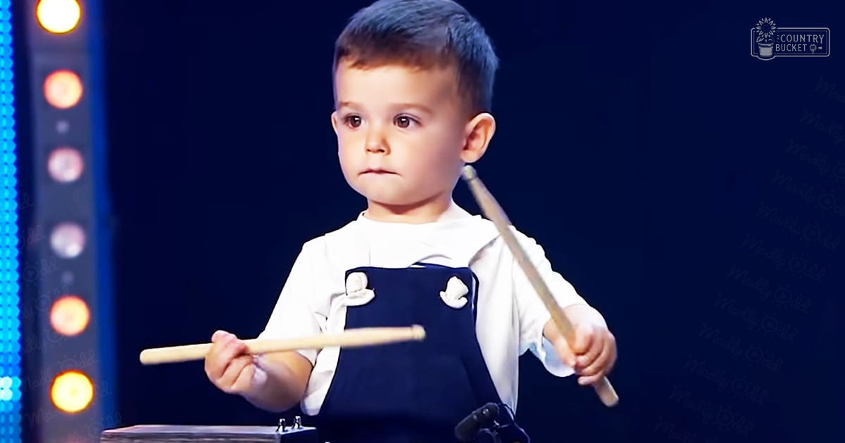 A toddler, approximately 3-4 years old, wearing a white long-sleeved shirt with a collar and dark blue overalls with white buttons, plays drums on stage. The setting is indoors with artificial lighting, suggesting an evening performance venue.