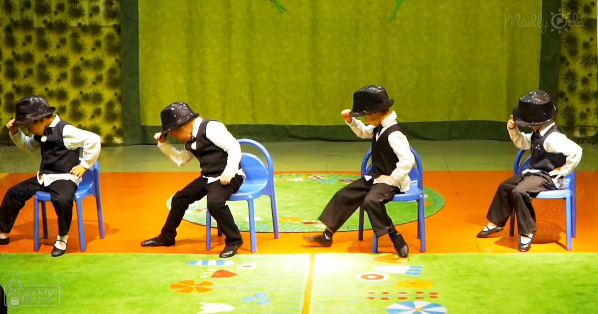 Four children in black vests, white shirts with rolled-up sleeves, black pants, and bowler hats perform on colorful mats against a green curtain resembling stylized trees or plants.
