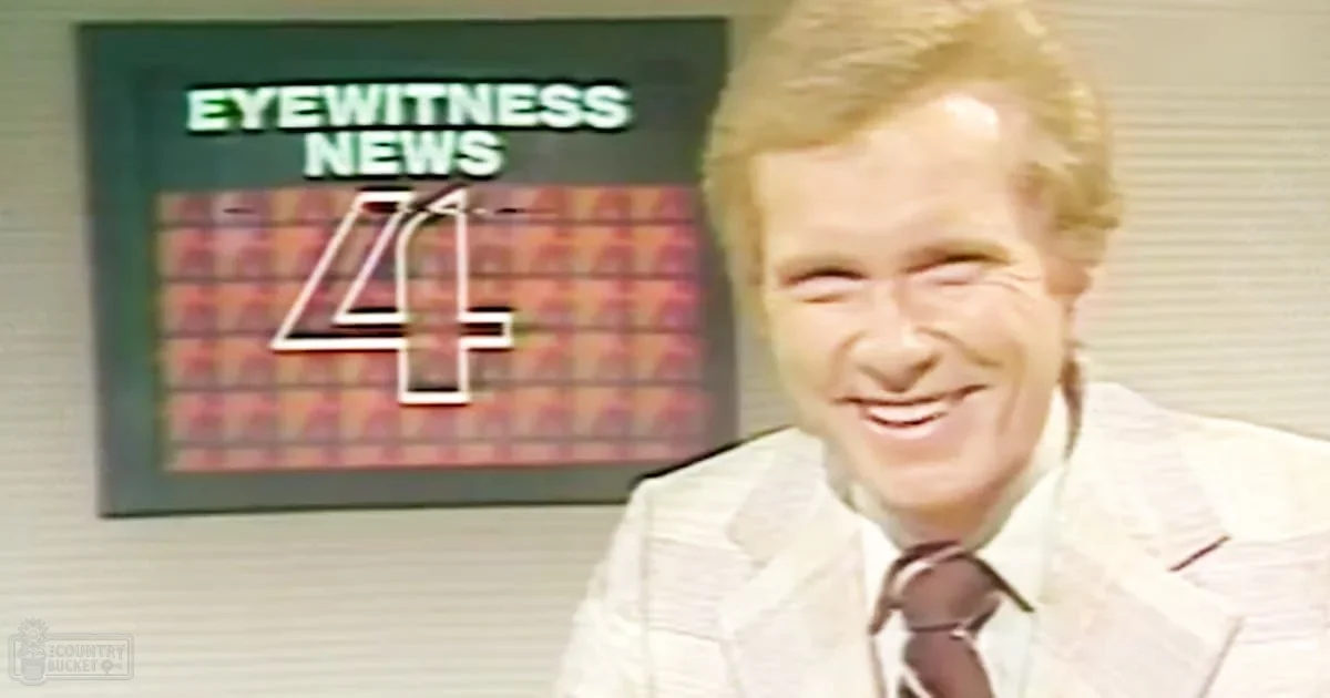 A middle-aged male news anchor in a light-colored suit jacket with a patterned design and dark tie stands in front of an 'Eyewitness News 4' sign, likely at a television studio. The setting is indoors with neutral-colored walls and bright, even lighting typical for professional studio environments.