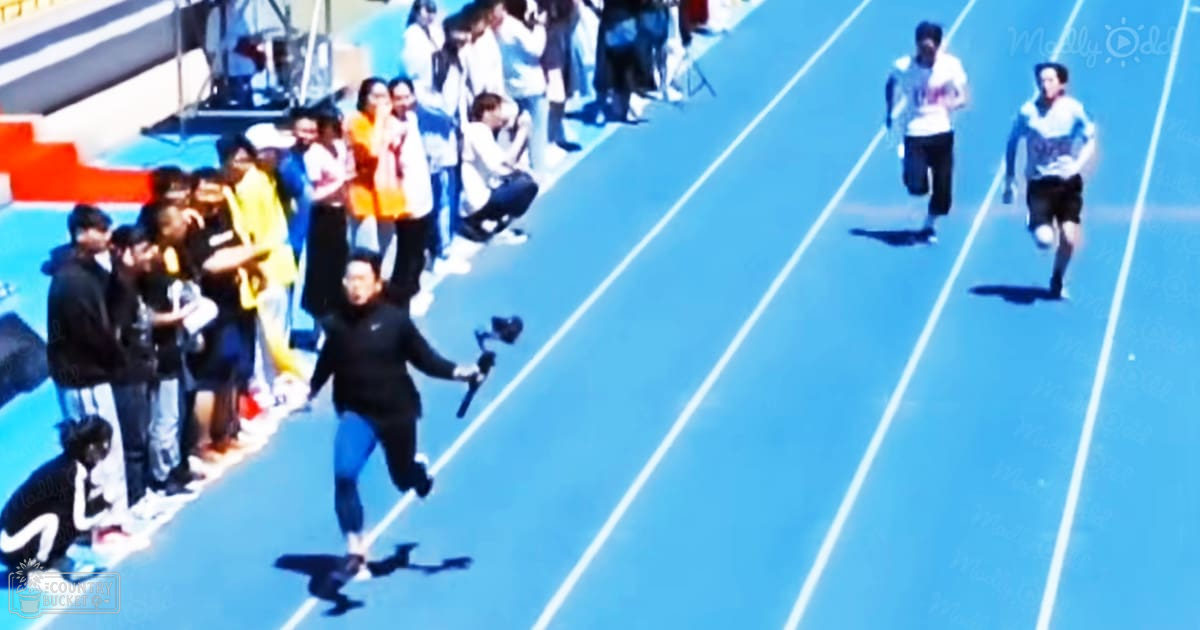 A young adult male running on a blue track with spectators watching at an outdoor sports stadium under clear daytime conditions.