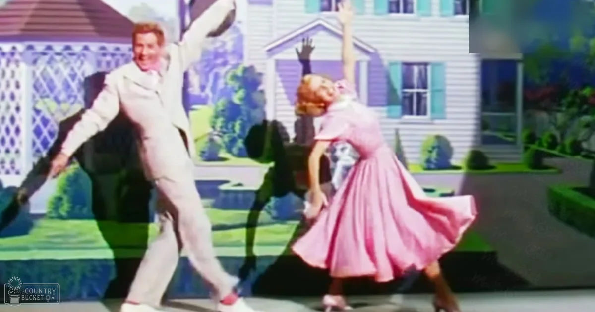 A musical film scene with a male in a white suit and red shoes, and a female in a pink dress, set against a backdrop of a house with purple shutters and greenery.