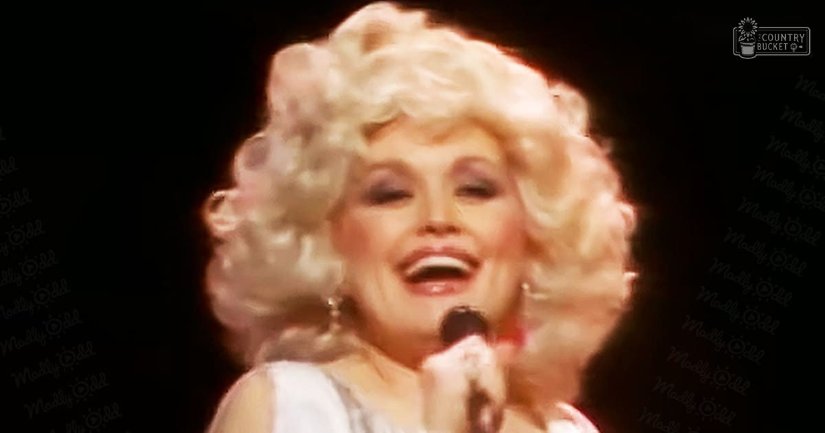 A middle-aged female performer in a concert setting wearing a shiny white satin or silk garment from the 1970s to early 1980s, holding a microphone under stage lighting.
