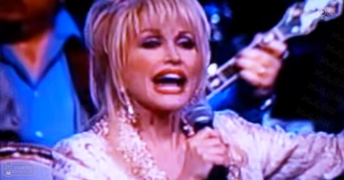 An adult female singer wearing an elegant lace top and large earrings performs on stage with dim lighting focused on her during a live concert.