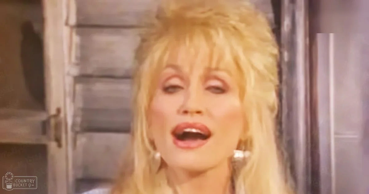 A middle-aged adult female singing indoors with large hoop earrings, light-colored clothing, and an old wooden door with shutters in the background suggesting a rustic setting.