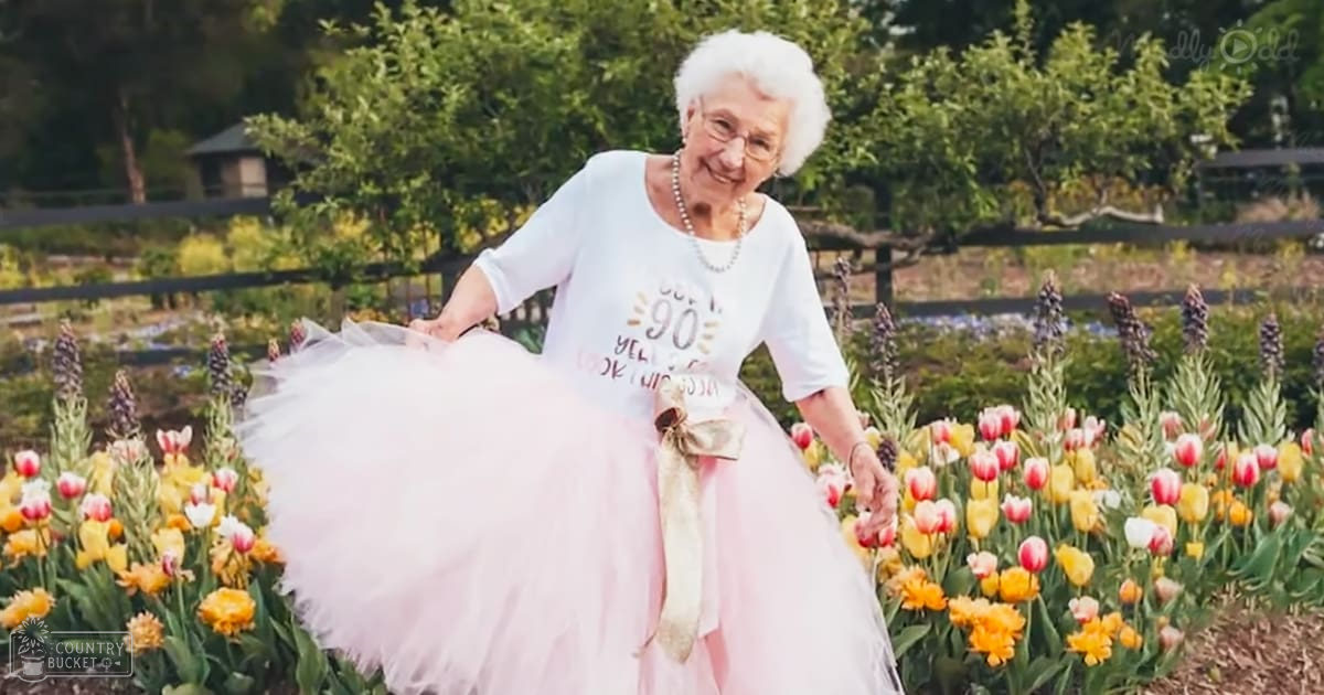 An older adult female in a garden setting, wearing a white t-shirt with text and decorative elements, a pink tutu skirt adorned with a gold ribbon, pearl necklaces, glasses, and possibly bracelets or rings, surrounded by greenery and tulips.