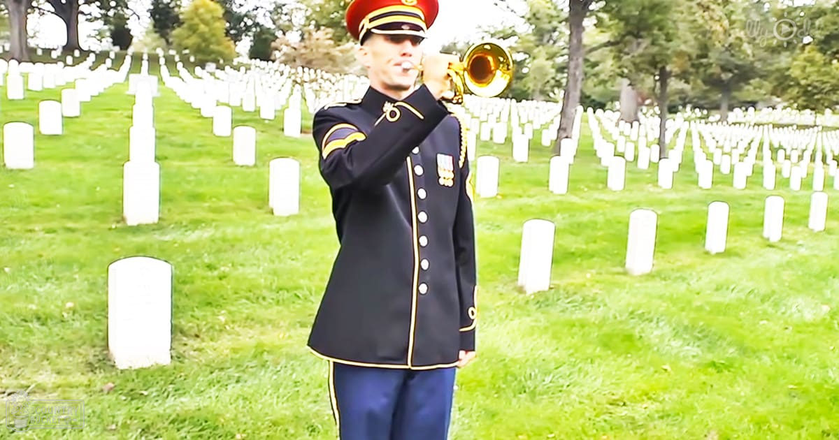 An adult male in a dark blue military uniform with gold trim and buttons plays the trumpet at Arlington National Cemetery during spring or summer under bright midday light.