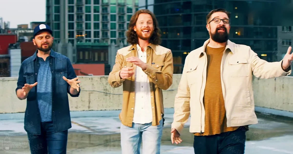 Three adult male singers perform outdoors on a rooftop, dressed casually with urban buildings in the background.