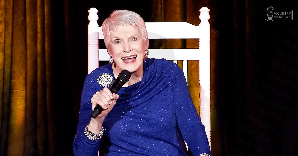 An older adult female performer wearing a glittery blue garment with beaded cuffs and large earrings stands on stage against a dark backdrop.