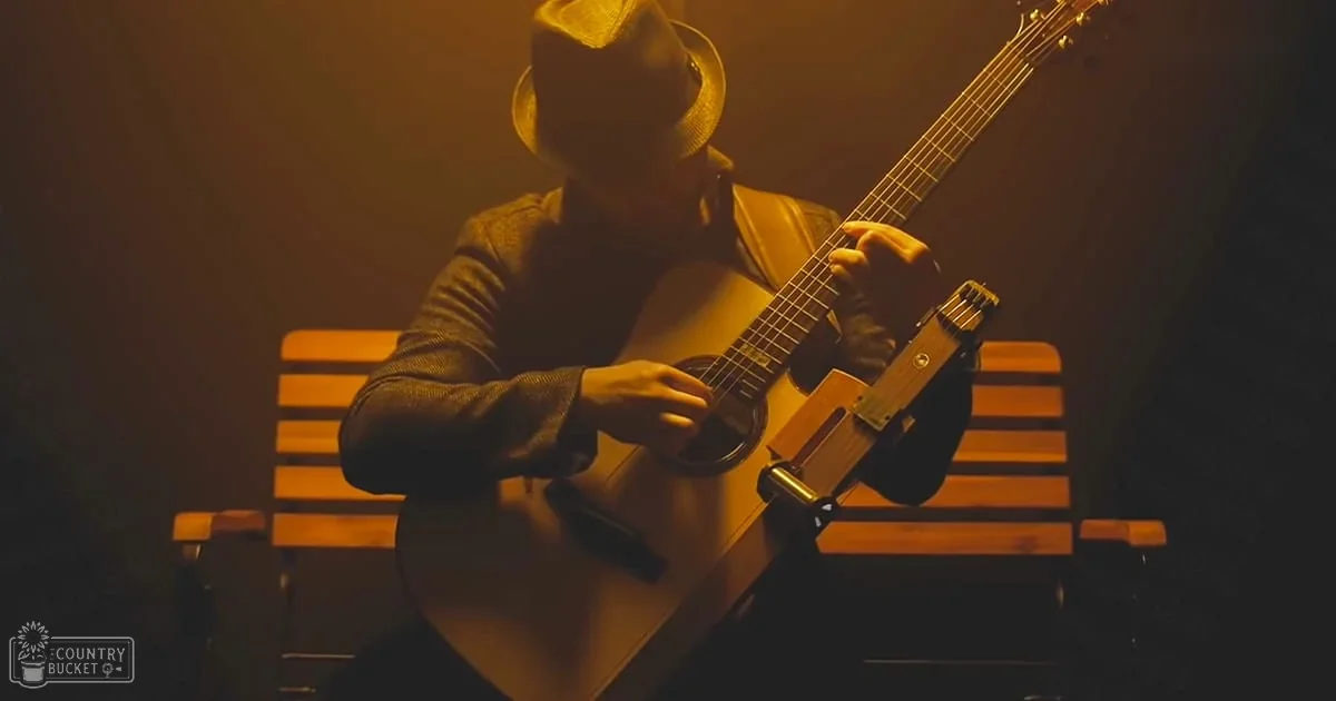 An adult male musician plays an acoustic guitar in a dimly lit indoor setting with warm yellowish-orange tones.