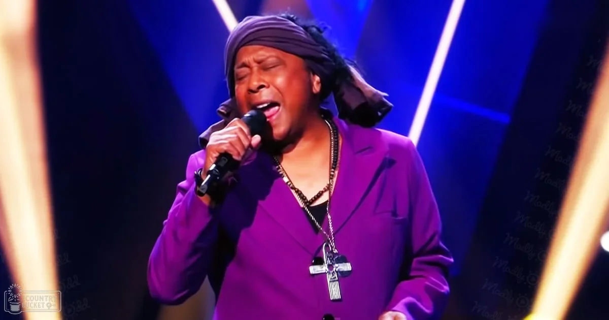 An adult male singer wearing a purple blazer, performing on an indoor stage with blue and white lighting.